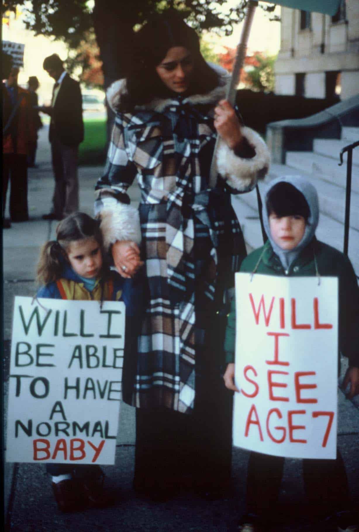 Image of mother and children. The children are holding signs that say "Will I be able to have a normal baby" and "Will I see age 7"